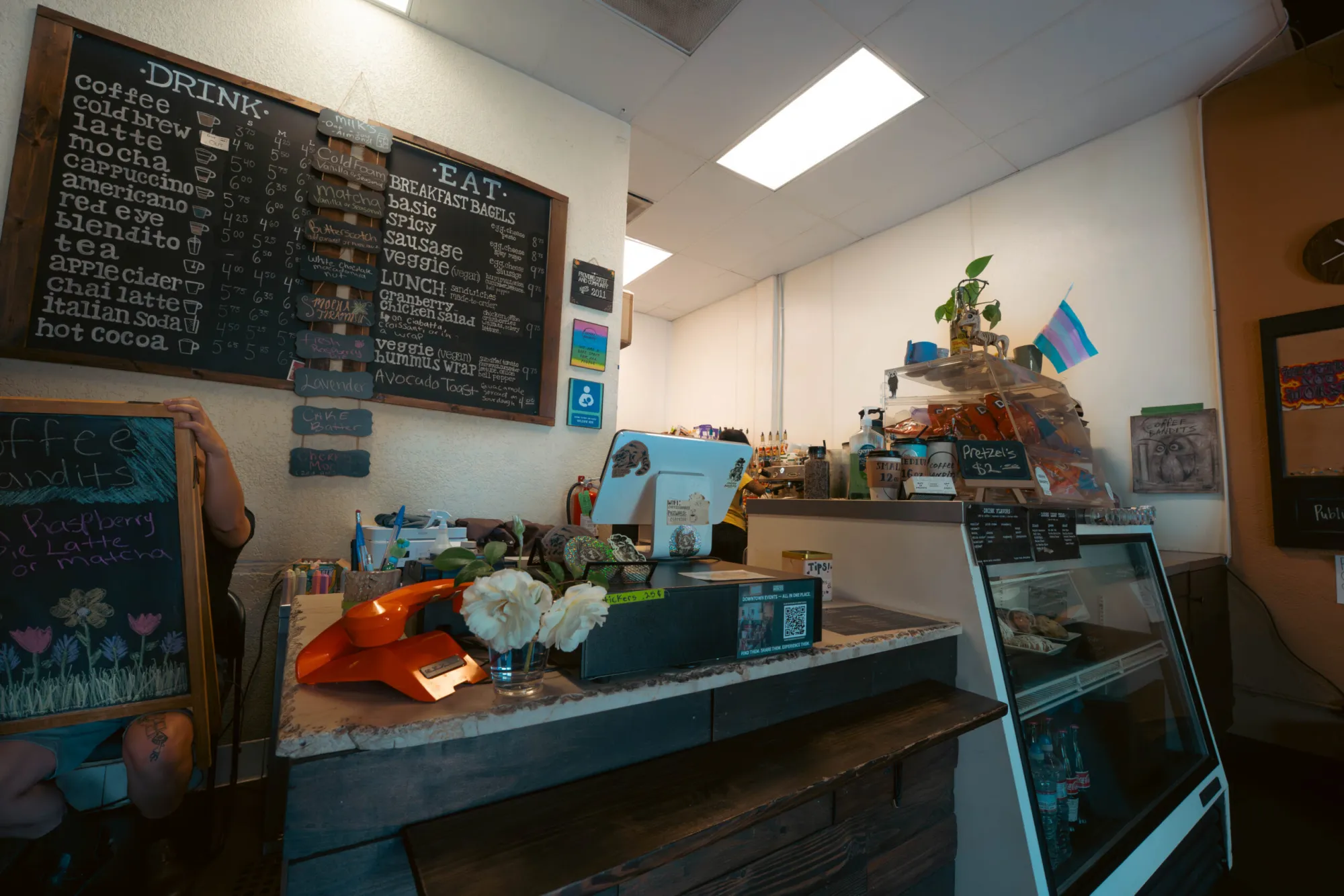 An indoor view of a coffee shop counter with an orange vintage telephone, a pastry display case, and a large chalkboard menu detailing drinks and breakfast items.