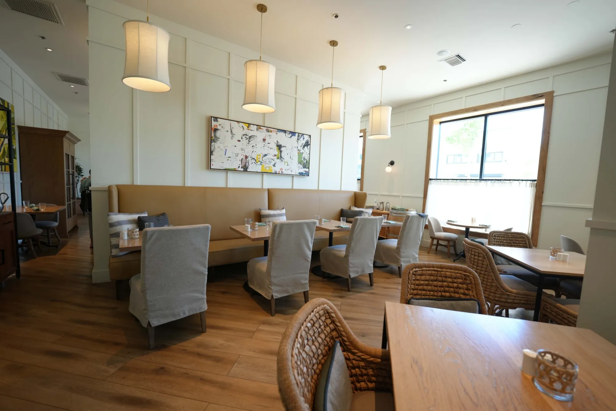 A bright and modern restaurant dining room featuring a long tan leather booth against a white paneled wall, light gray upholstered chairs, woven wicker seating, and contemporary pendant lights hanging over wooden tables.