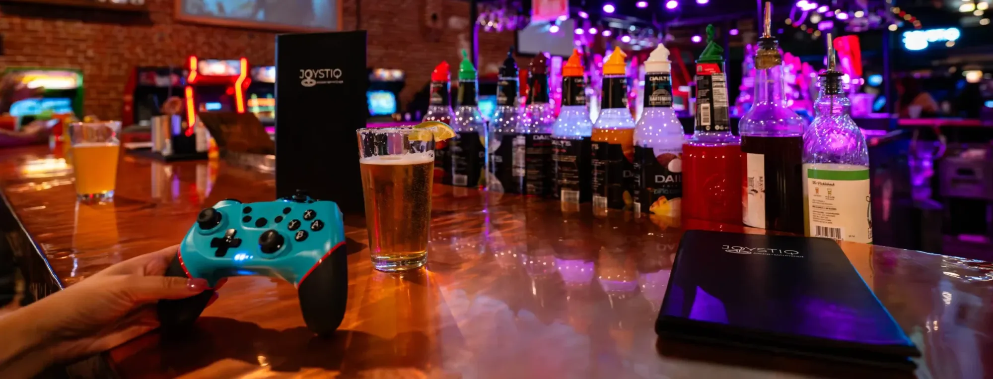 A hand holds a game controller at a bar counter lined with bottles, arcade machines behind