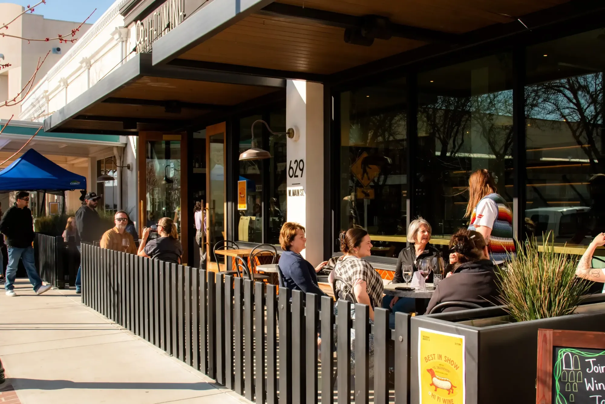 Outdoor patio seating at a storefront on W. Main St. with customers socializing on a sunny day.