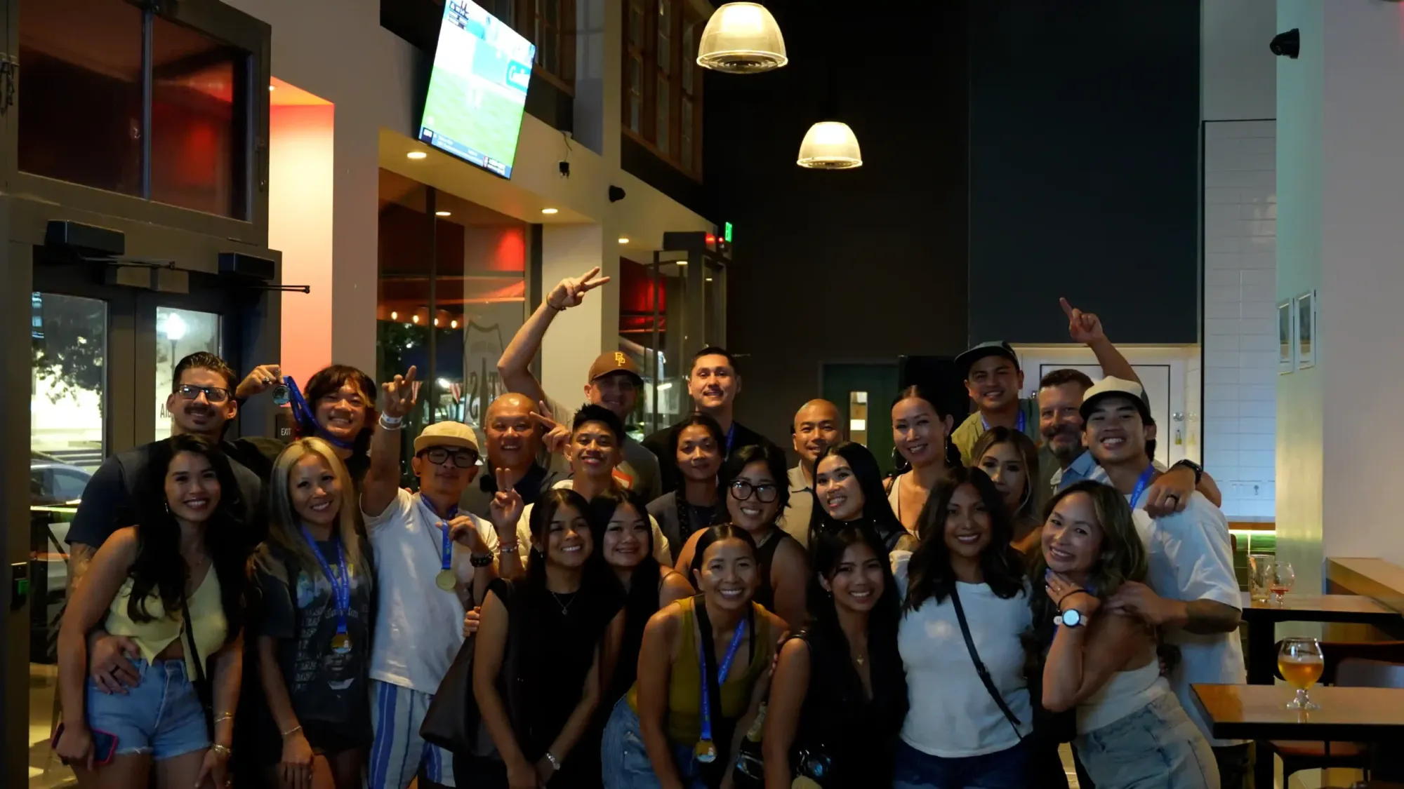 A large group of smiling friends posing for a celebratory photo indoors, with many individuals wearing medals or lanyards, and some visible interior decor including light fixtures and a television screen in the background.