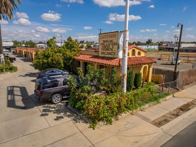 A high-angle shot of a retro single-story motel with a red tile roof and a tan exterior, featuring an old roadside sign reading "GATEWAY MOTEL" with a "VACANCY" notice, surrounded by green bushes and parked cars.
