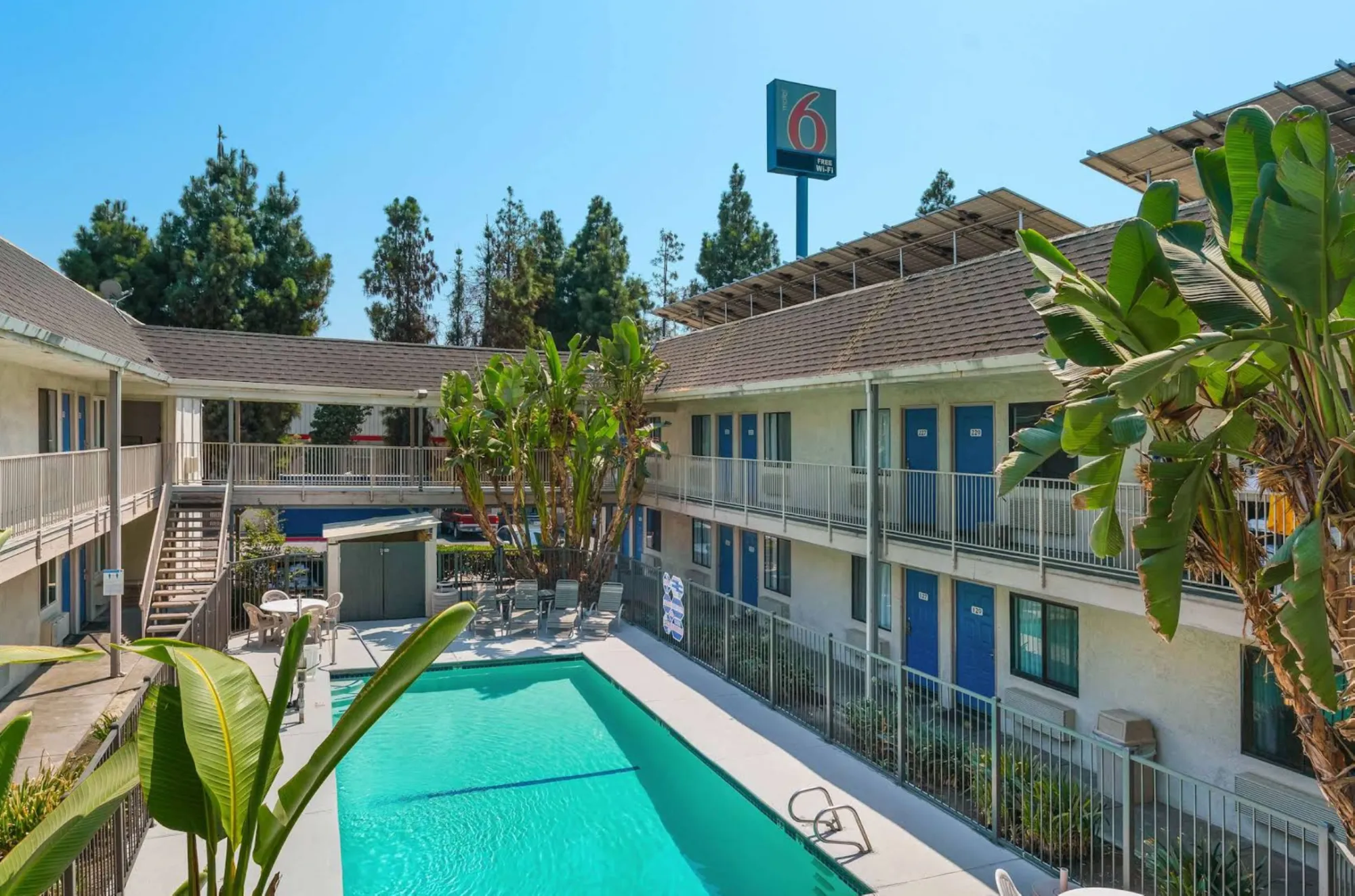A sunny courtyard of a two-story motel featuring an outdoor swimming pool filled with clear turquoise water, surrounded by metal fencing, concrete walkways, green palm trees, and a tall sign reading "Motel 6."