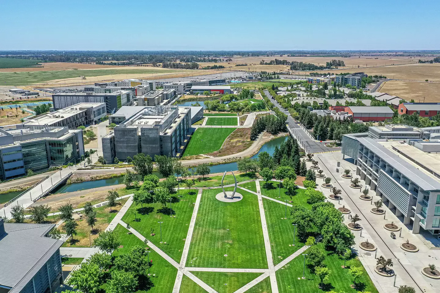 A high-angle aerial view of a modern university campus displaying structured green lawns, a winding canal, contemporary multi-story concrete academic buildings, and a large metallic arch sculpture in a central plaza.
