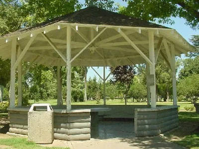 A large white wooden octagonal gazebo with a shingled roof sitting in a grassy public park surrounded by lush green trees on a sunny day.