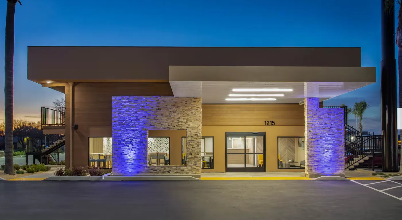 The exterior of a modern single-story hotel lobby at twilight, showcasing tan walls, decorative stone pillars illuminated with blue accent lights, large windows, and an overhanging entryway showing the address 1215.