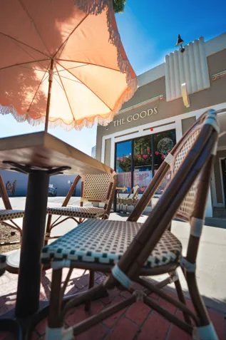 A low-angle, close-up shot of a woven bistro chair and patio table shaded by a large fringed pastel umbrella in front of "THE GOODS" boutique storefront.