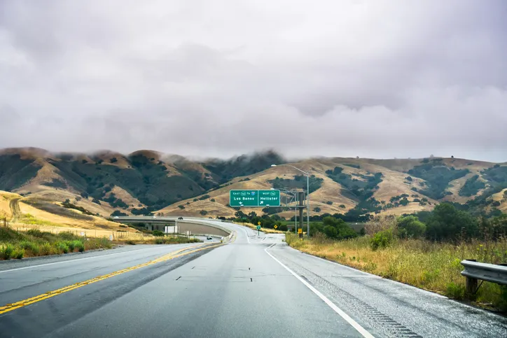 A wet asphalt highway stretching toward rolling green hills under an overcast sky, passing a green exit sign.