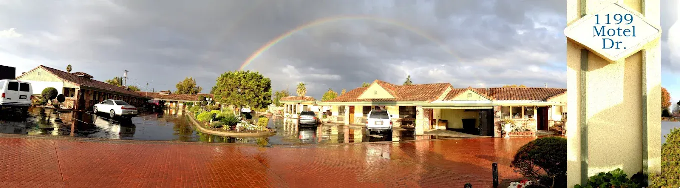 A wide panoramic view of a single-story motel with a wet asphalt parking lot and red brick driveway, featuring a vibrant rainbow arching across a dark, stormy sky above the buildings and a sign that reads "1199 Motel Dr."