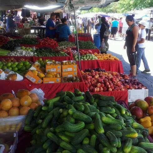 A crowded open-air market stall showcasing large piles of green cucumbers and oranges in the foreground with rows of other fresh fruits and vegetables on red-clothed tables.