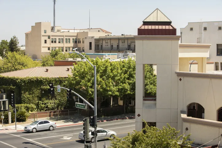 An elevated view of a city street intersection with a sign reading "18th Street," featuring silver cars driving past modern tan commercial buildings and lush green trees on a sunny day.