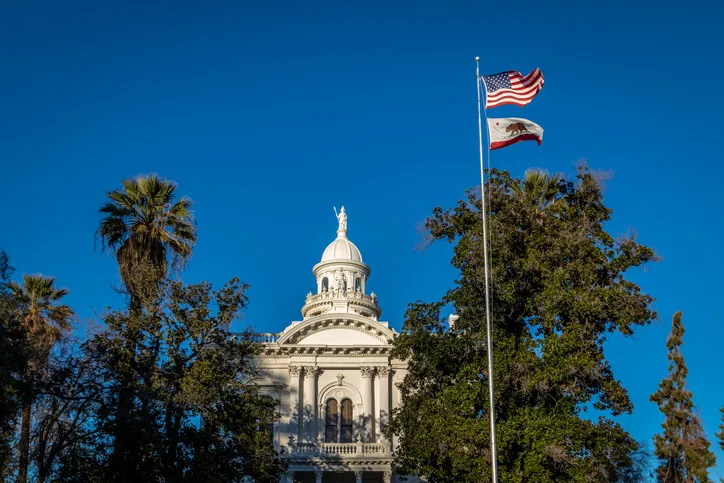 The historic white dome and classical architecture of a courthouse building rising above green trees, with the United States flag and the California Republic flag flying together on a tall flagpole under a clear blue sky.