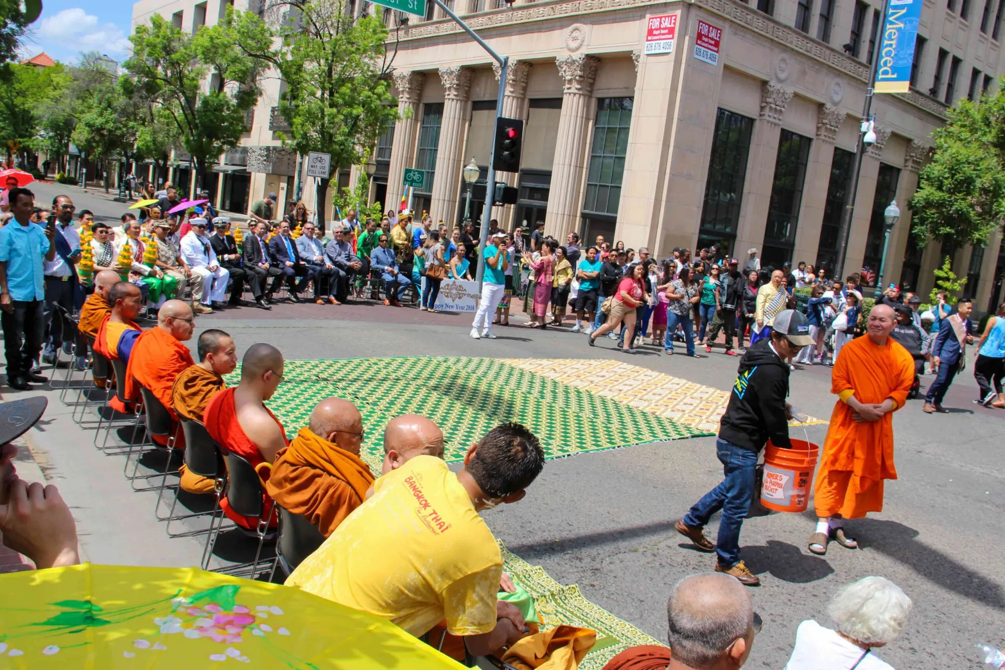 Buddhist monks in vibrant orange robes participate in an outdoor street parade surrounded by community spectators.