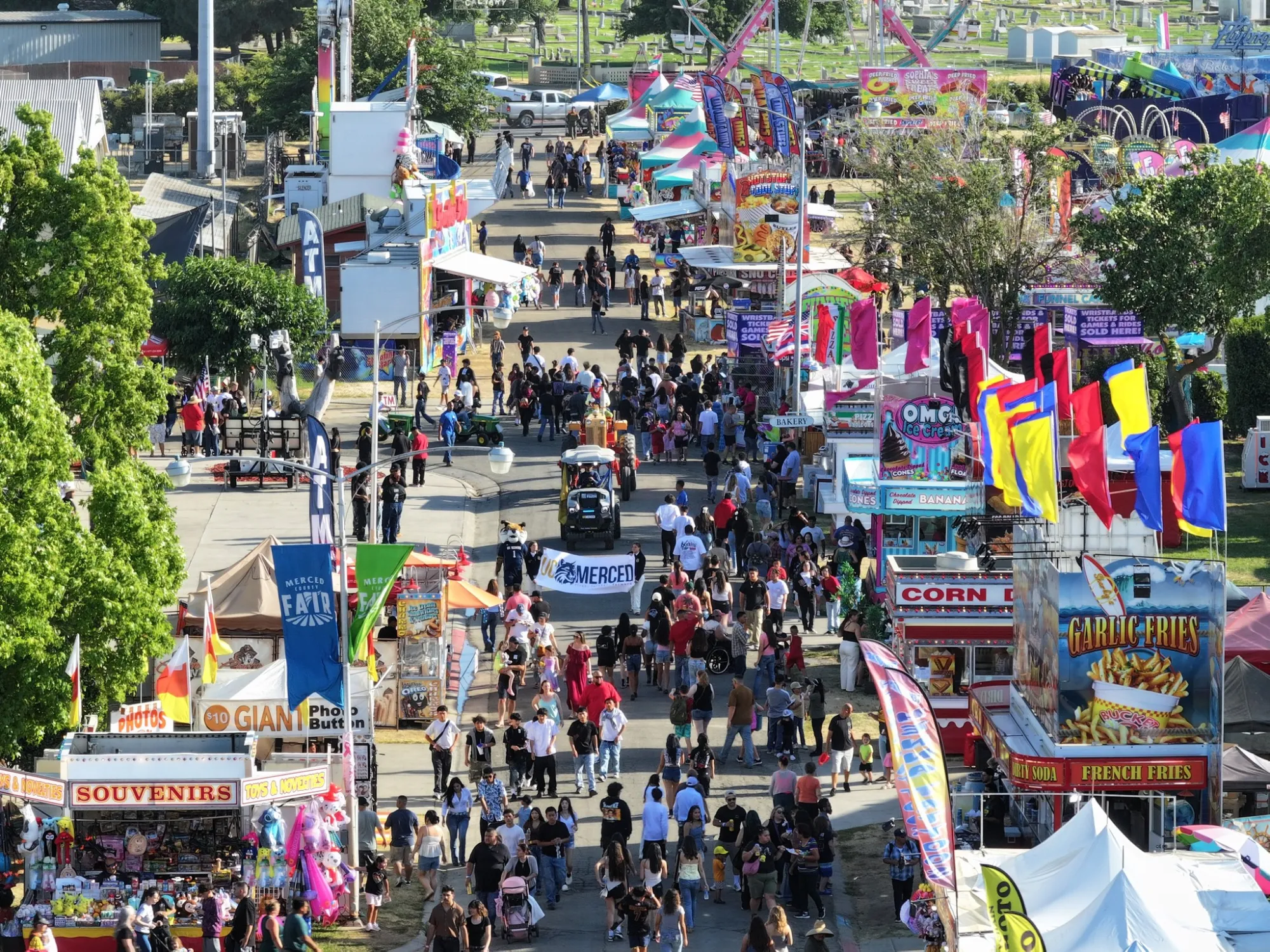 A bustling, high-angle view of a crowded street fair midway filled with colorful food booths selling garlic fries and ice cream, with a "UC Merced" banner visible in the crowd.