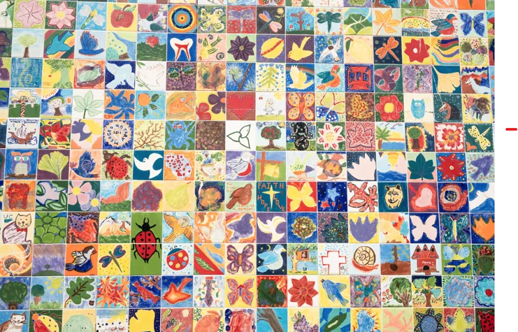 A colorful public art mural composed of dozens of small, hand-painted ceramic tiles featuring diverse drawings of animals, flowers, birds, and community themes.