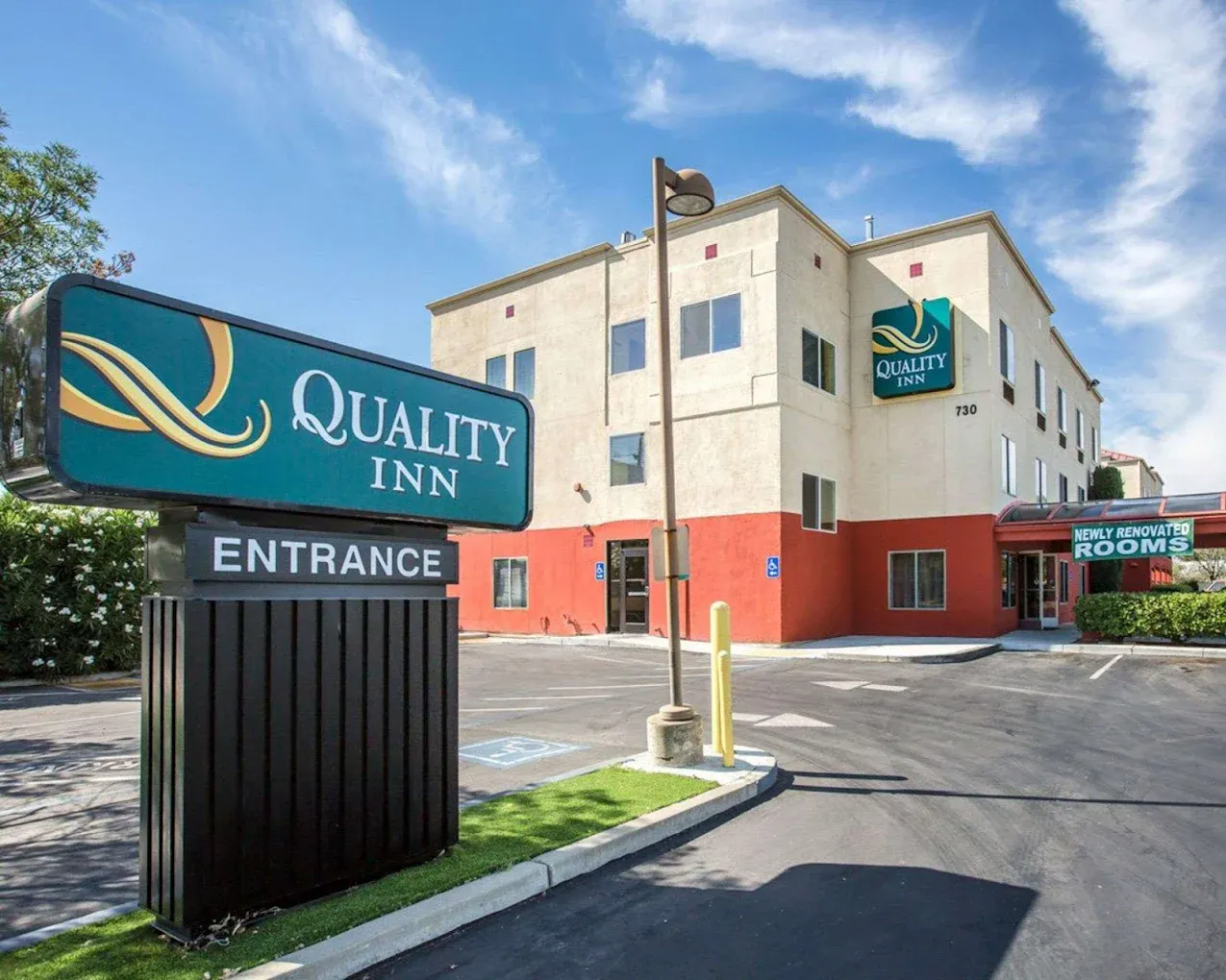 A large teal and white sign reading "QUALITY INN ENTRANCE" stands in front of a three-story beige and red hotel building under a bright blue sky with wispy white clouds.