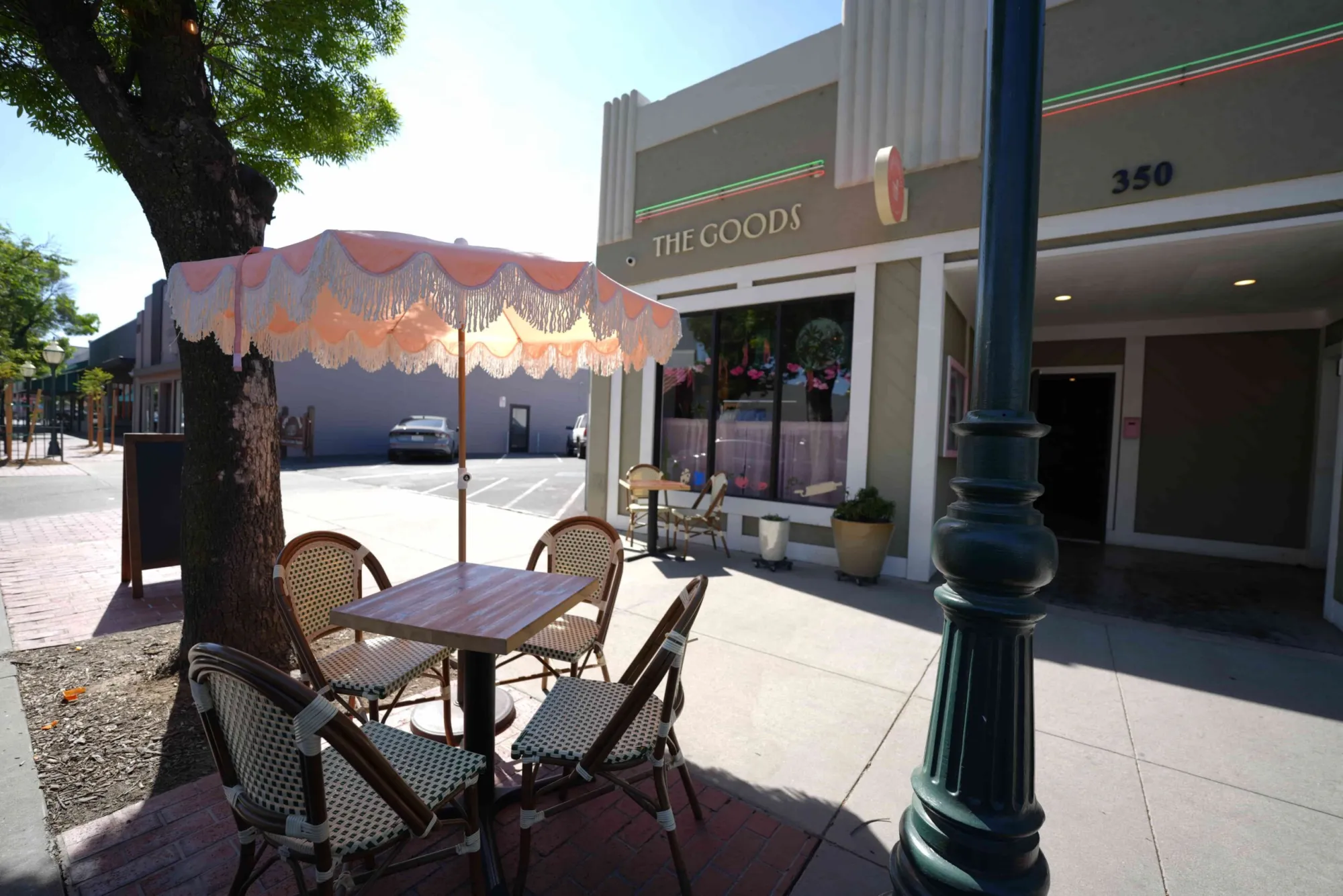 An outdoor patio dining table with a fringed pastel umbrella sitting on a brick sidewalk in front of a boutique shop named "The Goods."