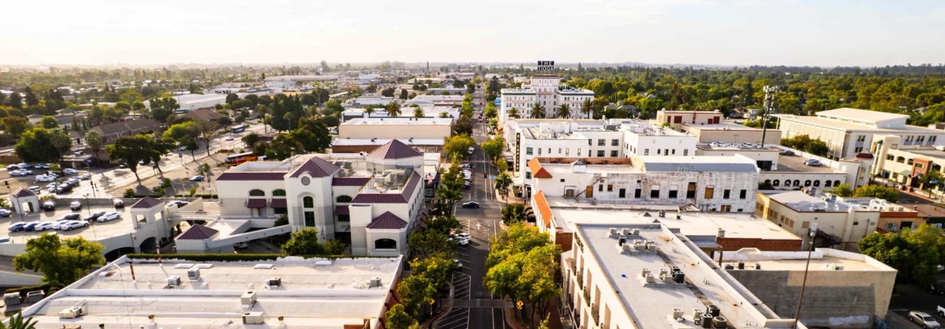 Aerial drone view of downtown Merced streets lined with historic buildings, palm trees, and parked cars under a bright sky.