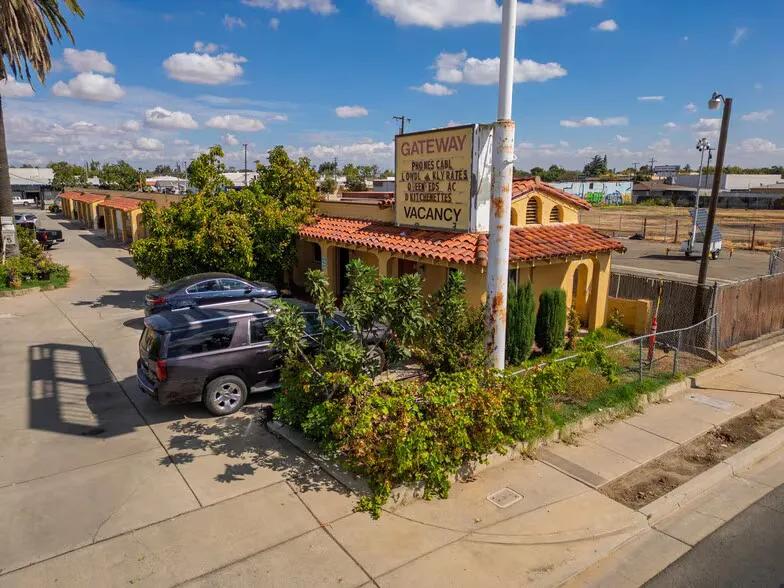 A high-angle shot of a retro single-story motel with a red tile roof and a tan exterior, featuring an old roadside sign reading "GATEWAY MOTEL" with a "VACANCY" notice, surrounded by green bushes and parked cars.