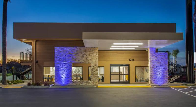 The exterior of a modern single-story hotel lobby at twilight, showcasing tan walls, decorative stone pillars illuminated with blue accent lights, large windows, and an overhanging entryway showing the address 1215.