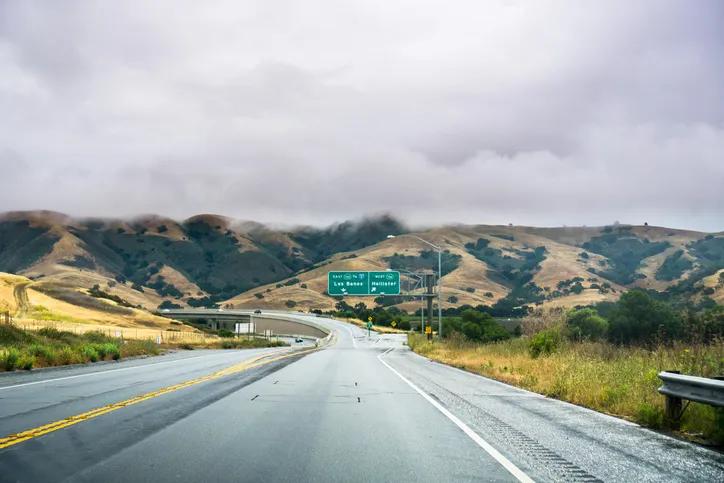 A wet asphalt highway stretching toward rolling green hills under an overcast sky, passing a green exit sign.