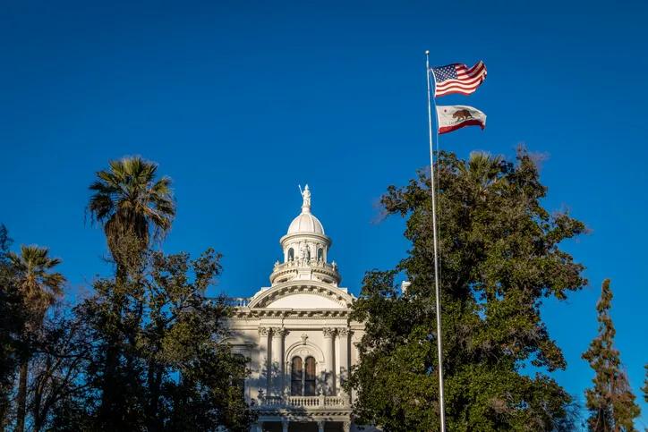 The historic white dome and classical architecture of a courthouse building rising above green trees, with the United States flag and the California Republic flag flying together on a tall flagpole under a clear blue sky.